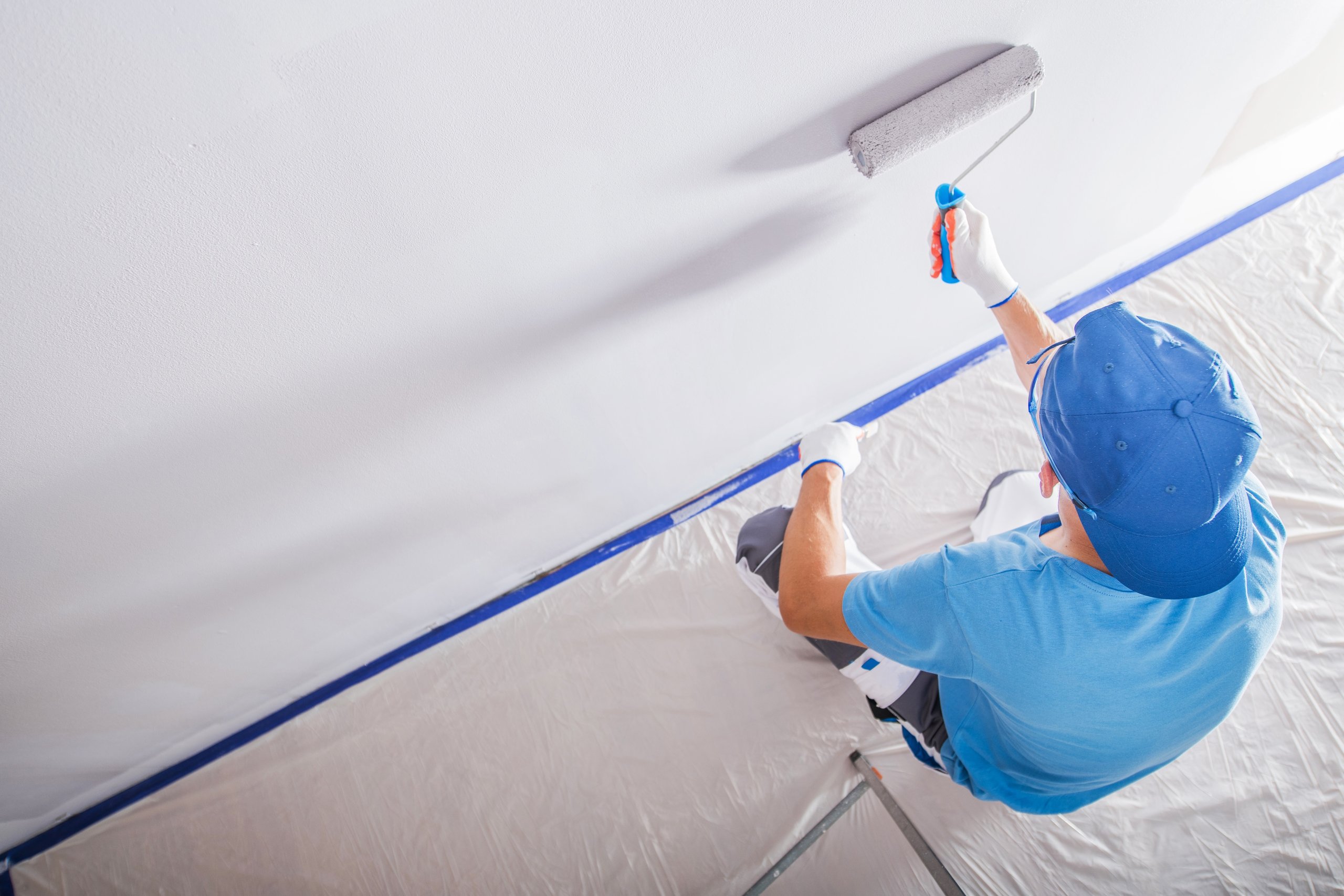 Painter wearing blue cap and shirt applying white paint to a wall with a roller while sitting on a step stool.