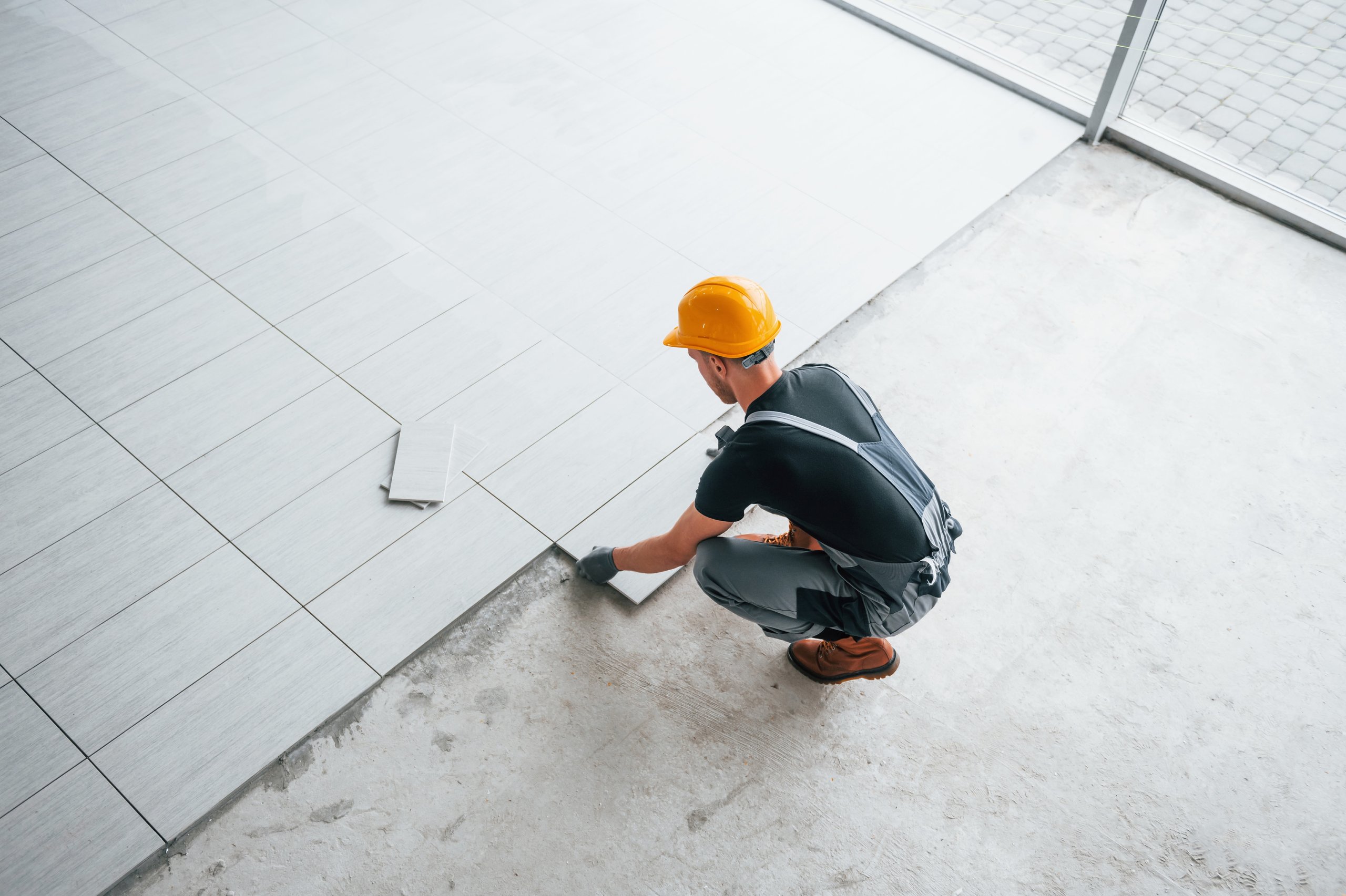 Worker wearing a yellow hard hat and gloves installs large light gray floor tiles over a concrete surface near a window.