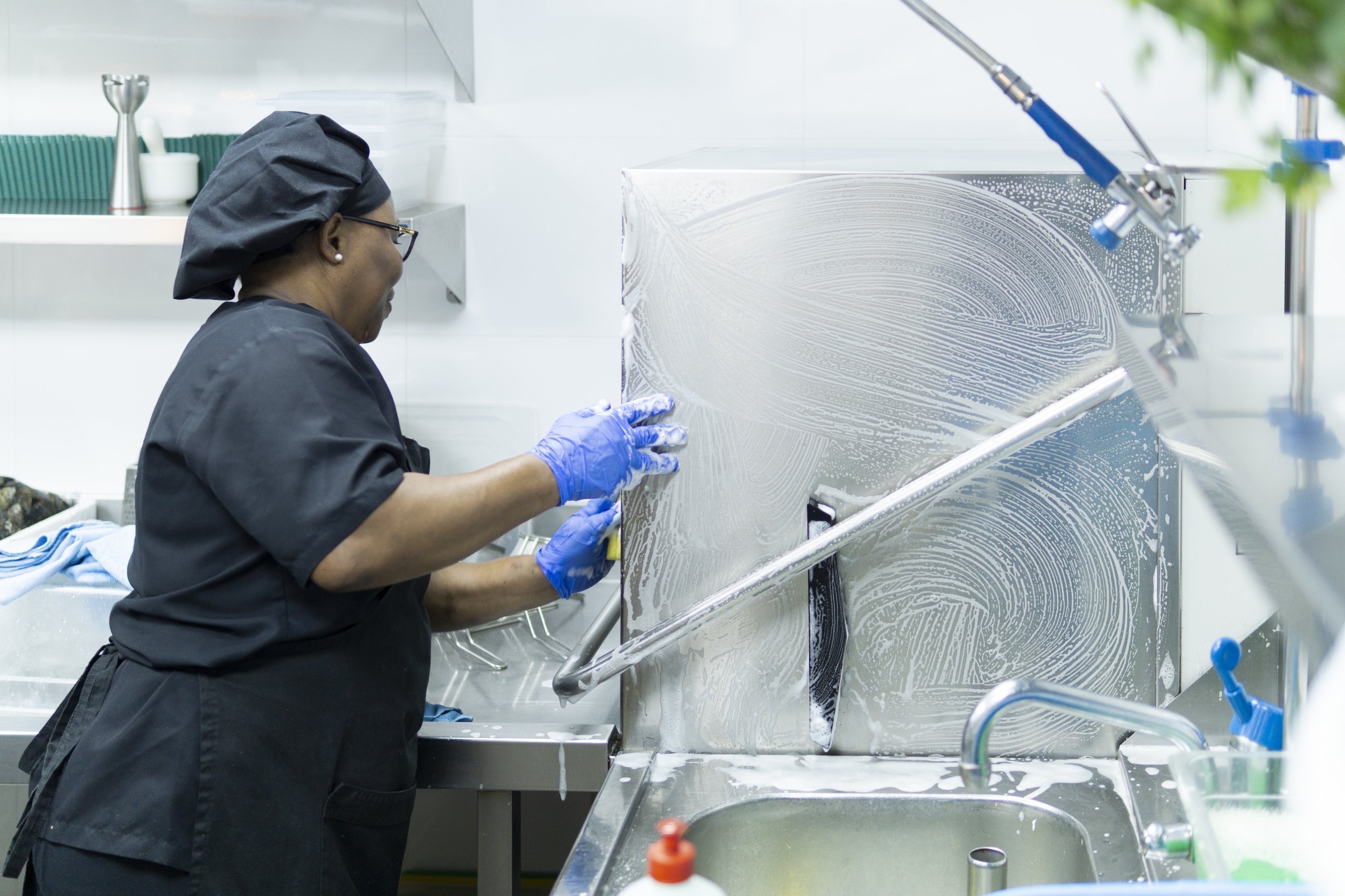 Woman in black uniform and blue gloves washing a large, soapy industrial kitchen dishwasher.