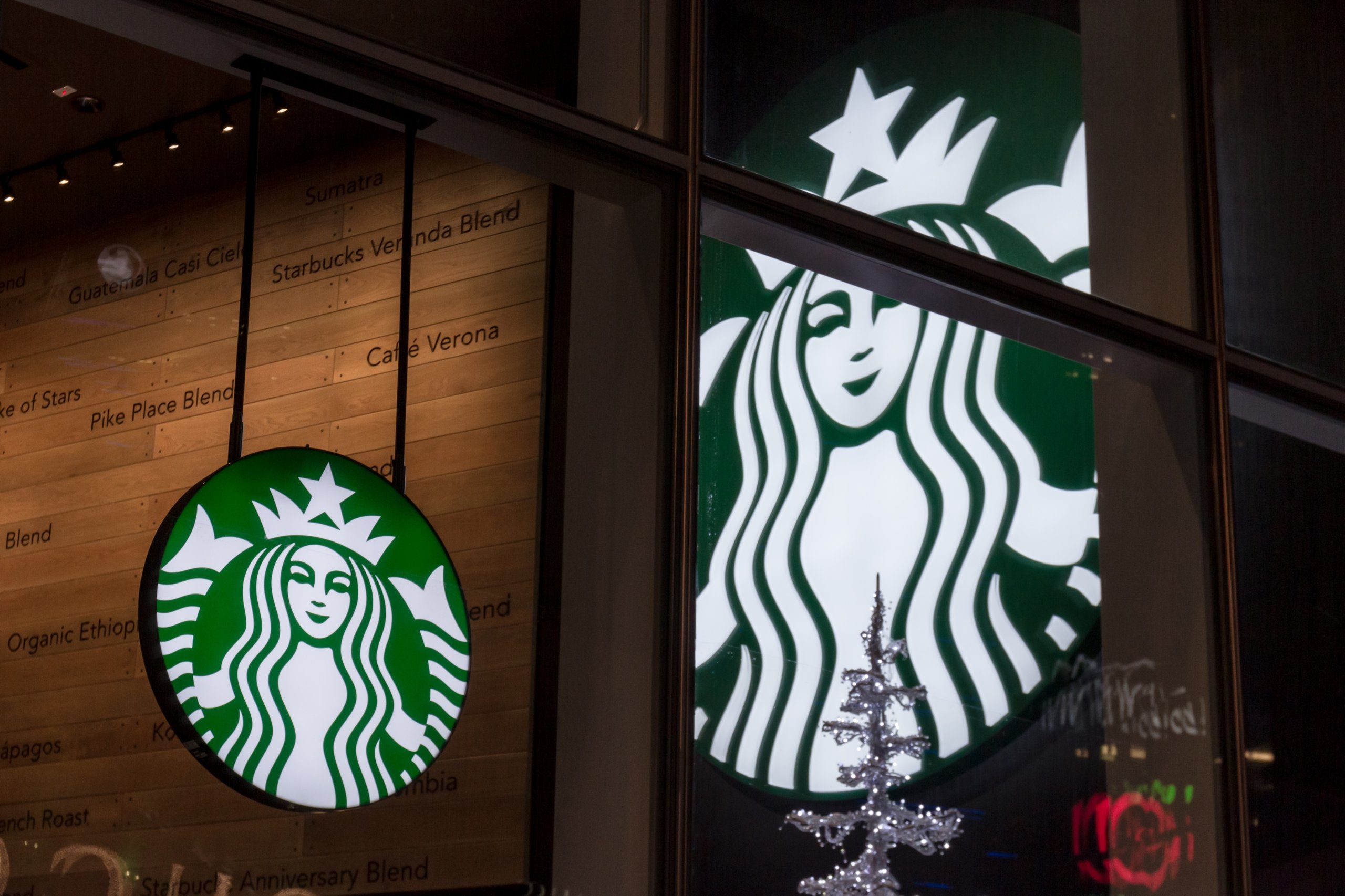 Illuminated Starbucks logo sign hanging inside a coffee shop, reflected on a glass window at night.
