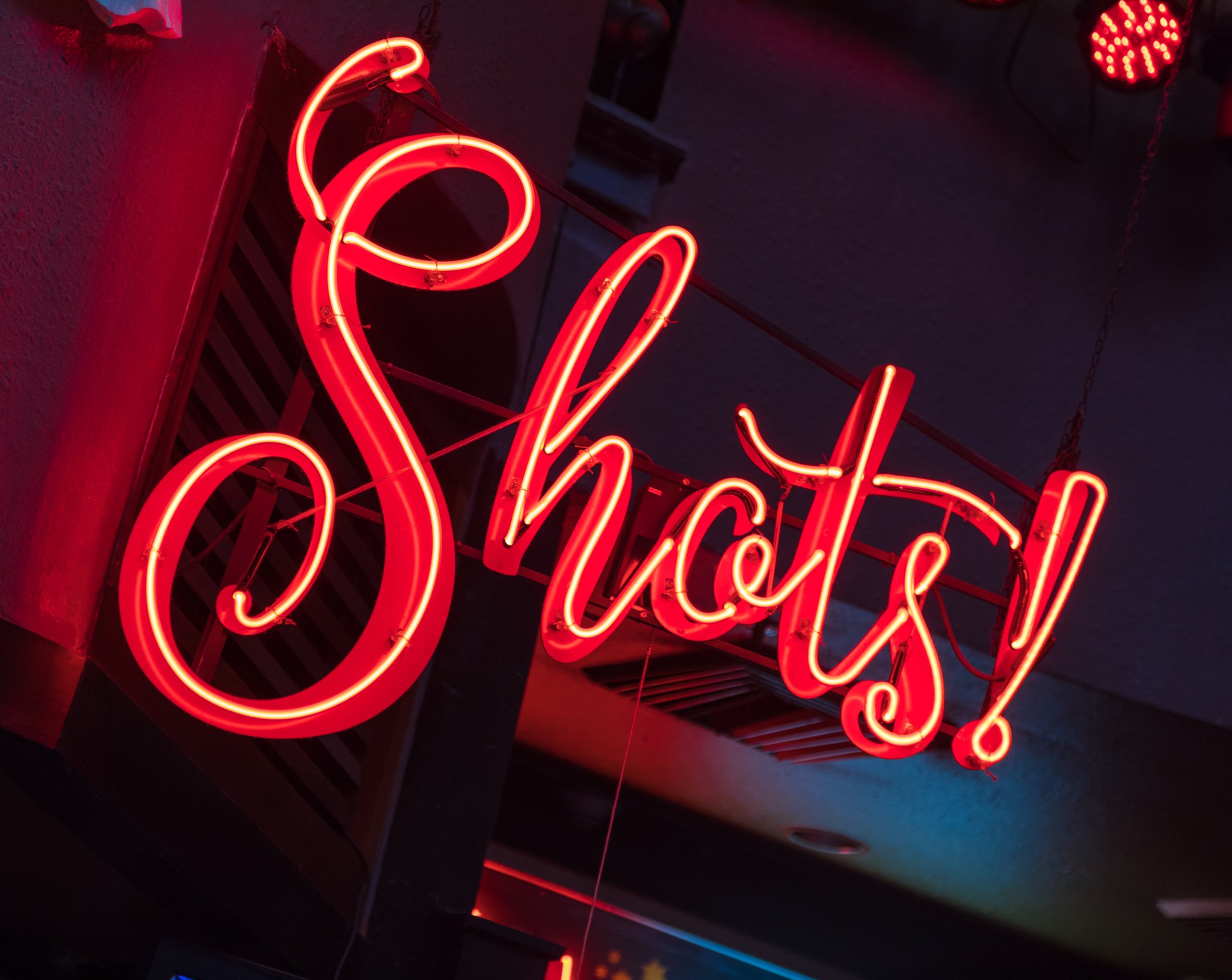 Bright red neon sign displaying the word 'Shots!' hanging indoors above a bar area.