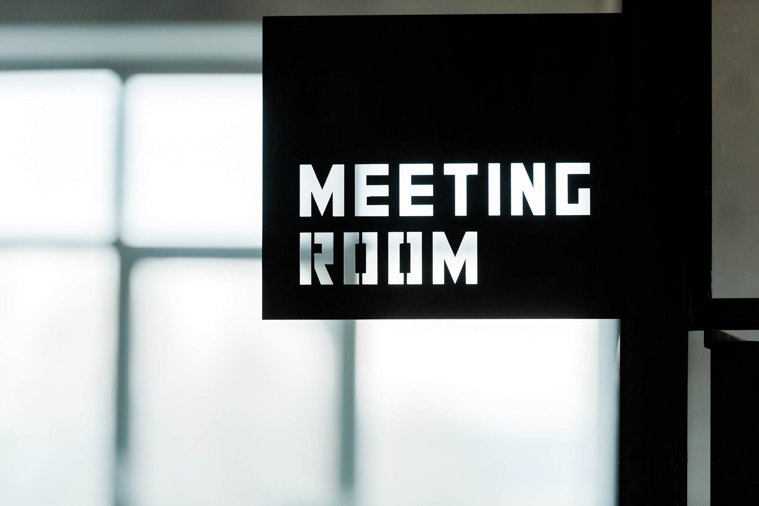 Black sign with white text reading 'MEETING ROOM' mounted on a wall near a window.