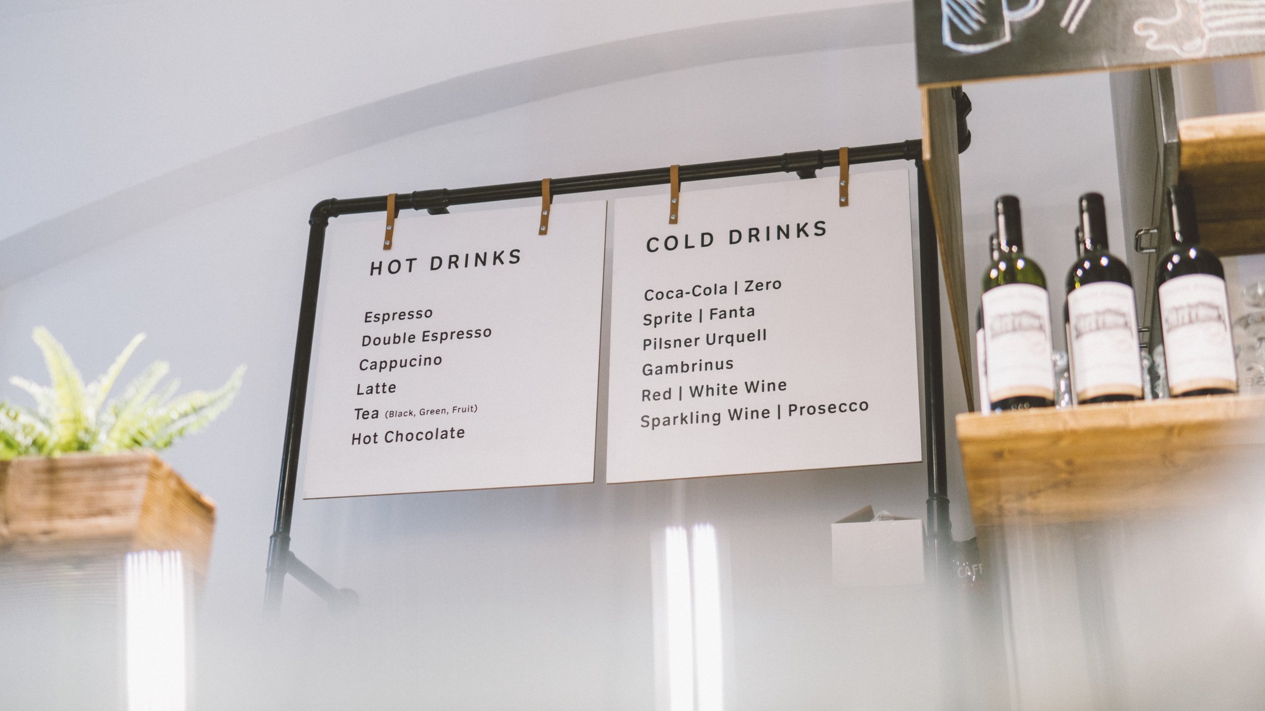 Cafe menu board listing hot drinks including espresso, cappuccino, latte, tea, hot chocolate, and cold drinks such as Coca-Cola, Sprite, pilsner, and wine.