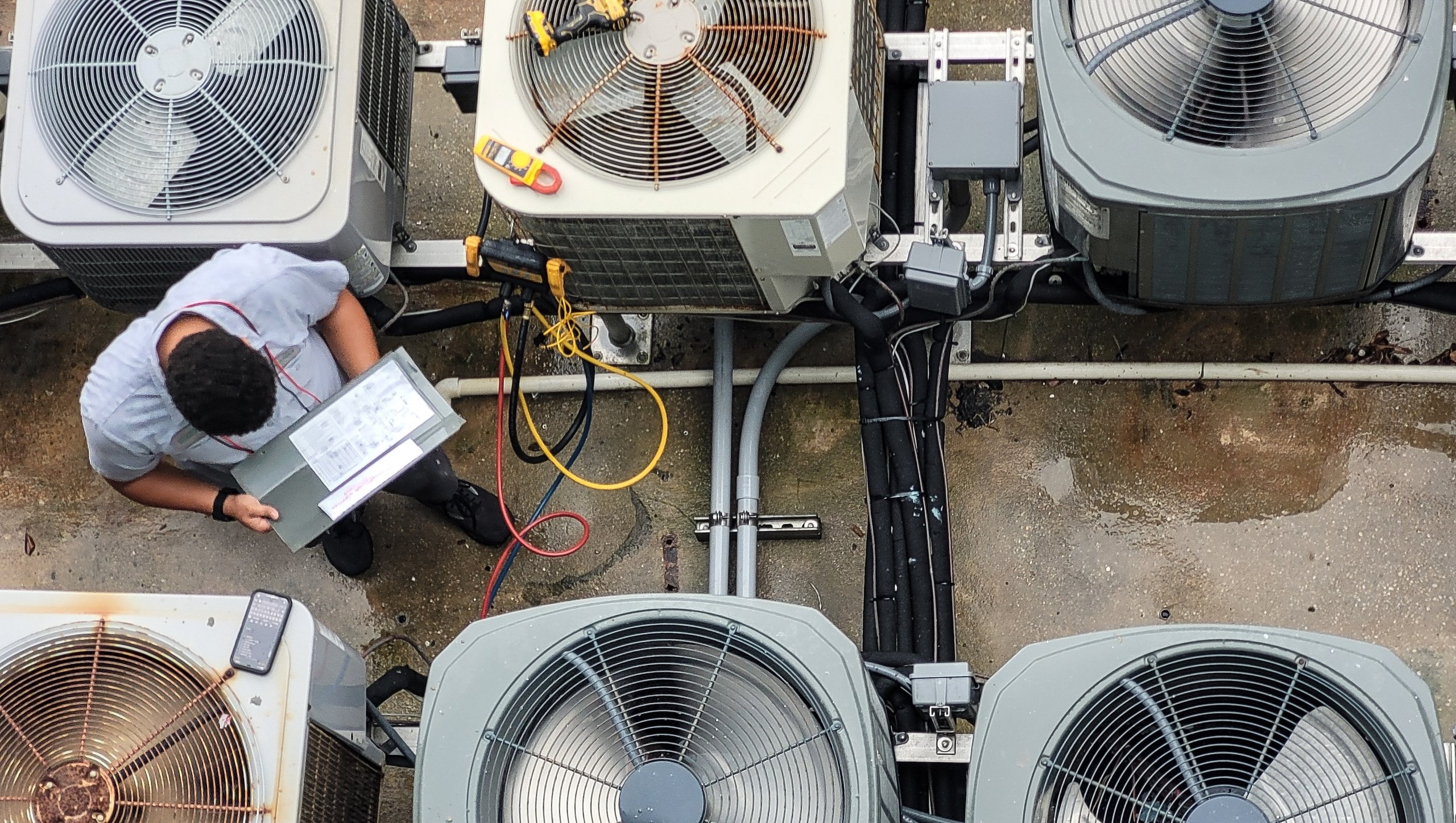 Technician inspecting and holding a control panel box among multiple outdoor air conditioning units with visible wiring and pipes on a concrete surface.
