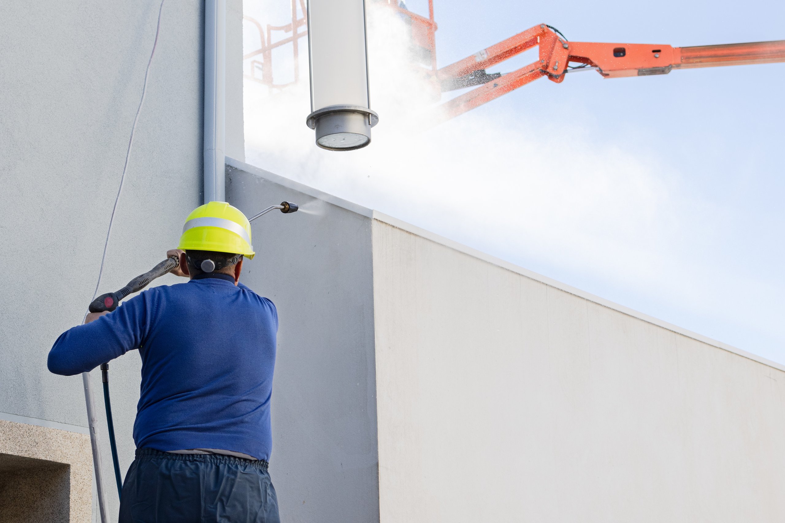 Worker wearing a yellow hard hat pressure washing the side of a grey building wall.