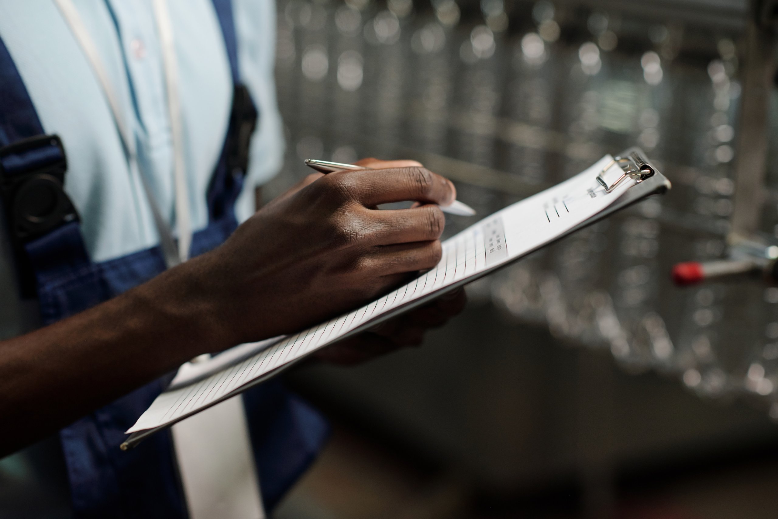 Close-up of a person in work attire writing on a clipboard with a pen in an industrial or warehouse setting.