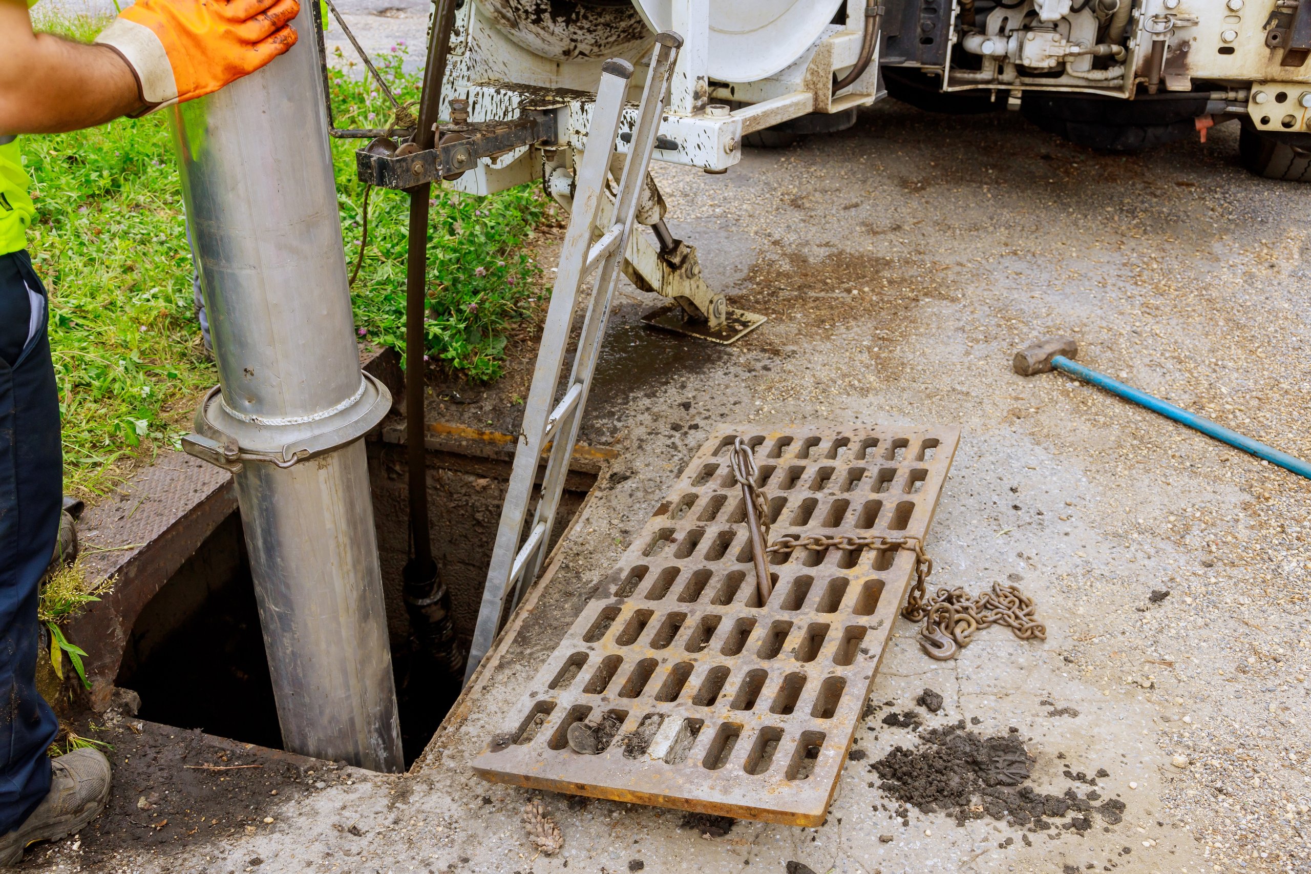 Worker wearing orange gloves operating a large metal pipe into an open sewer manhole beside a ladder and a lifted sewer grate chained to a rod.