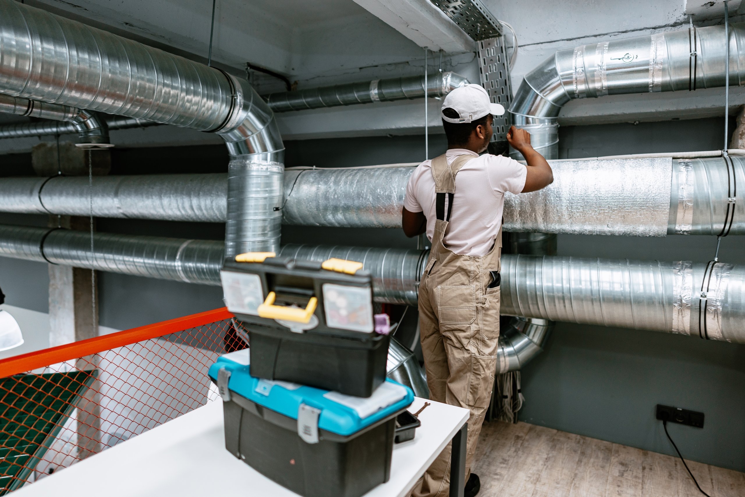 Technician in beige overalls and white cap working on large silver HVAC ducts in an industrial setting.