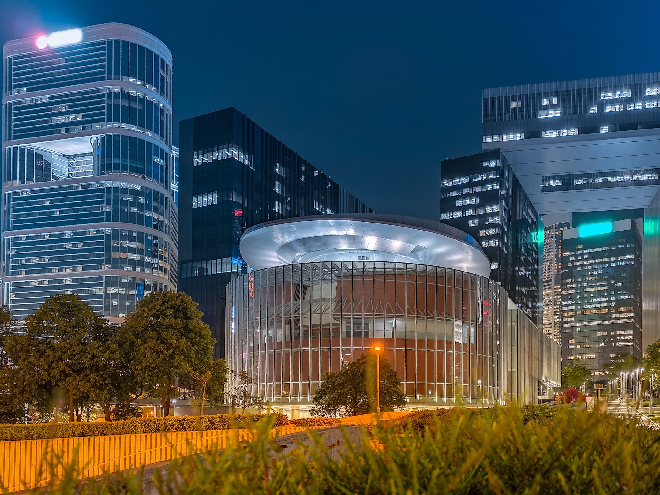 Modern cityscape at night featuring illuminated high-rise buildings and a circular glass structure in the foreground.