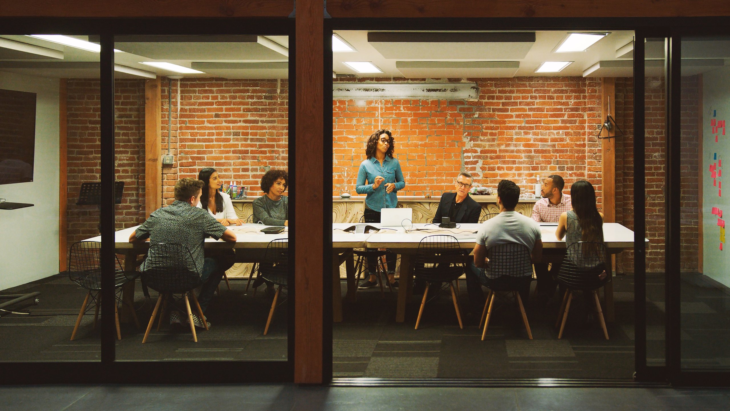 Employees sitting around a white conference table with one woman standing and speaking in a modern office with brick walls.