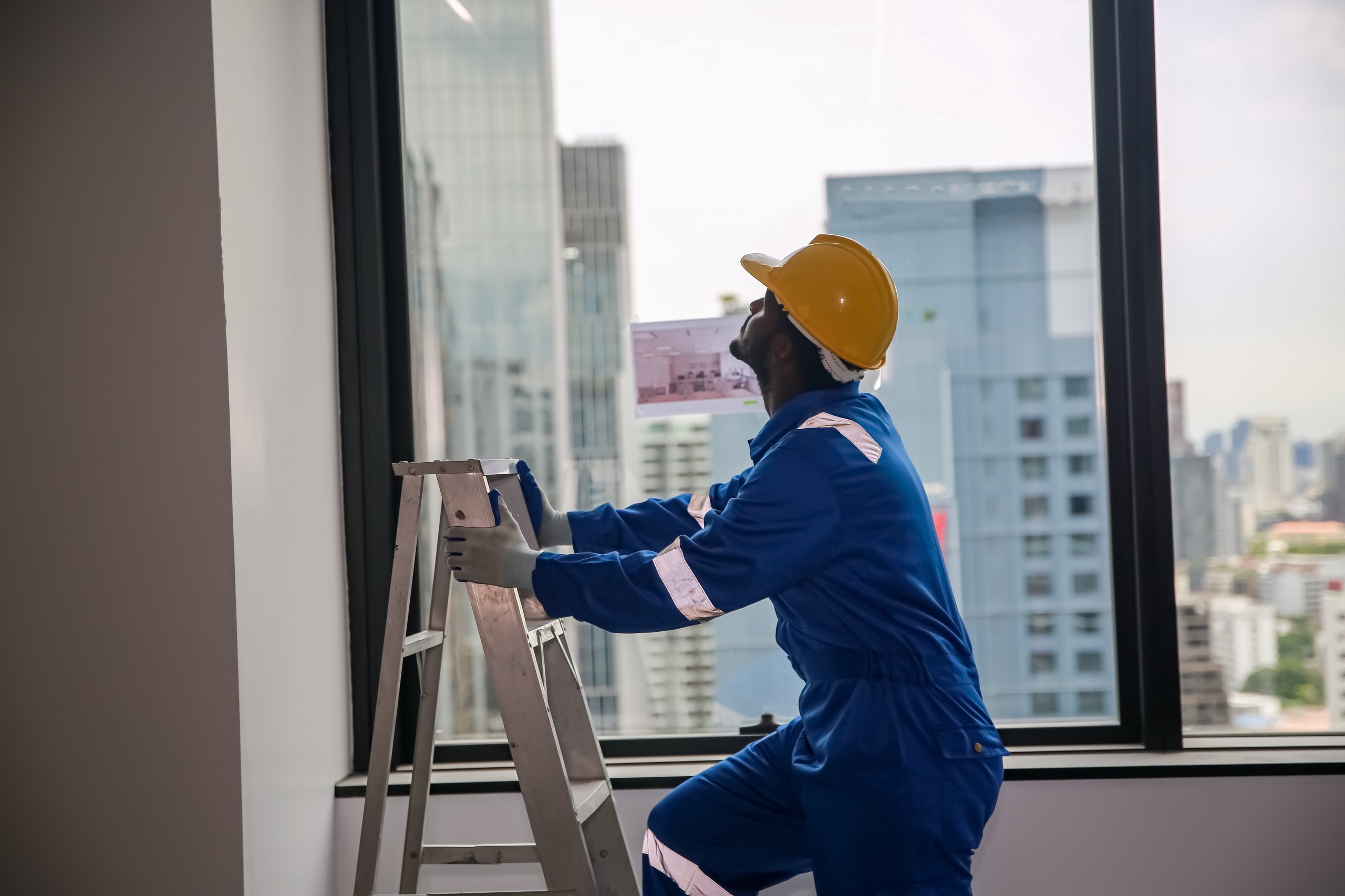 Worker in blue protective suit and yellow hard hat inspecting a metal ladder near a large window overlooking a cityscape.