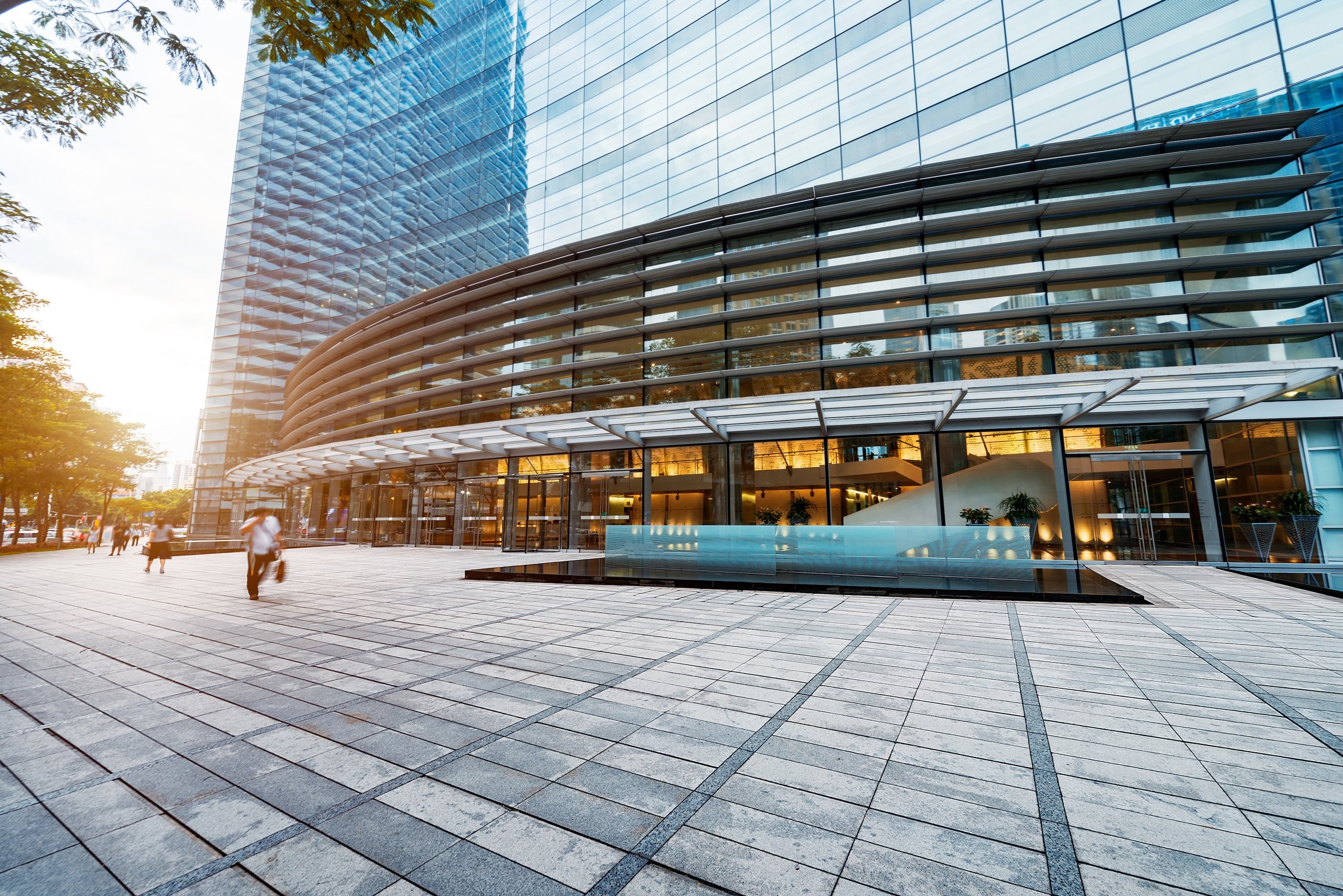Modern glass office building with curved facade and people walking on the spacious tiled plaza at sunset.