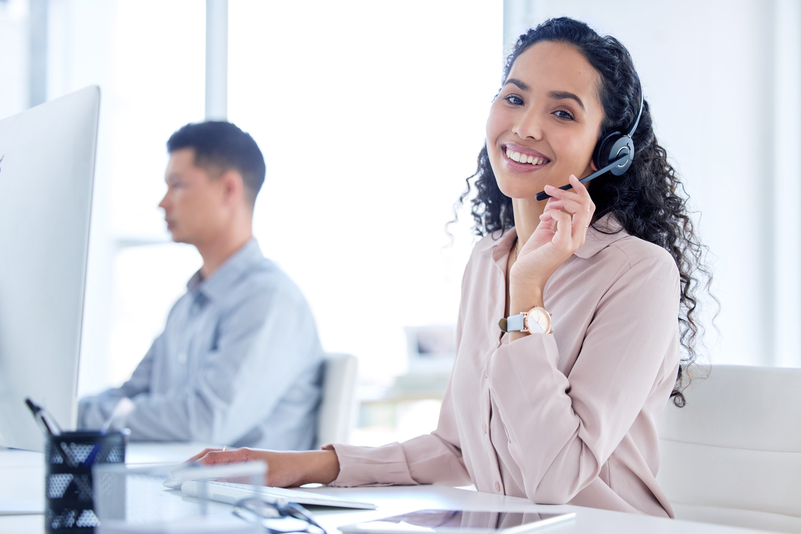 Smiling woman with curly hair wearing a headset, working at a computer in a bright office.