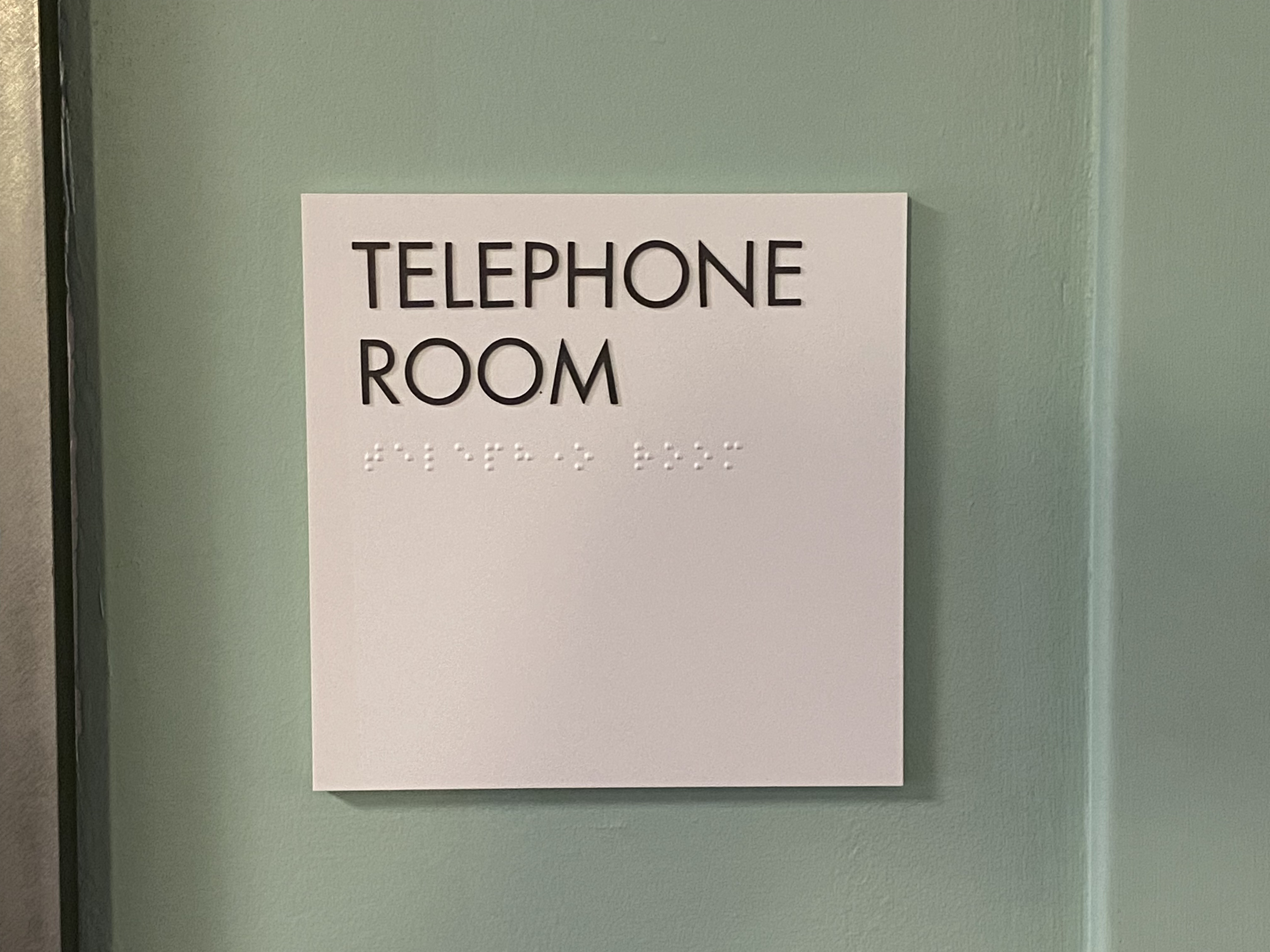 White wall sign with black text reading 'TELEPHONE ROOM' and Braille below, mounted on a green wall.