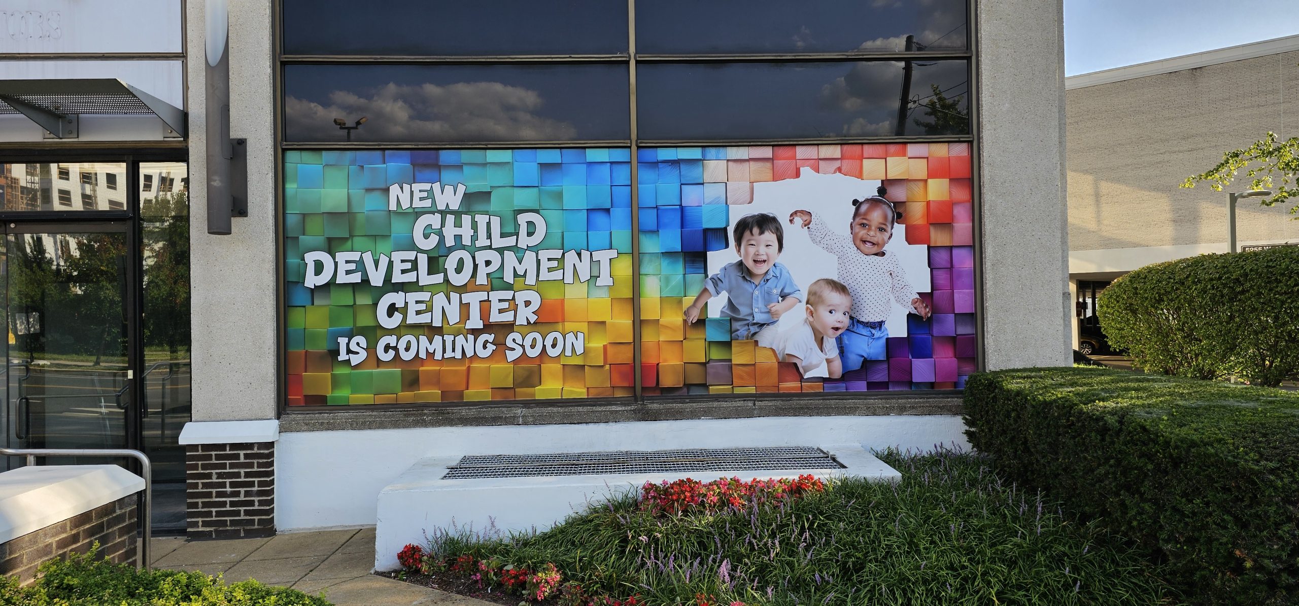Window mural announcing a new child development center coming soon, featuring three diverse toddlers smiling against a colorful mosaic background.