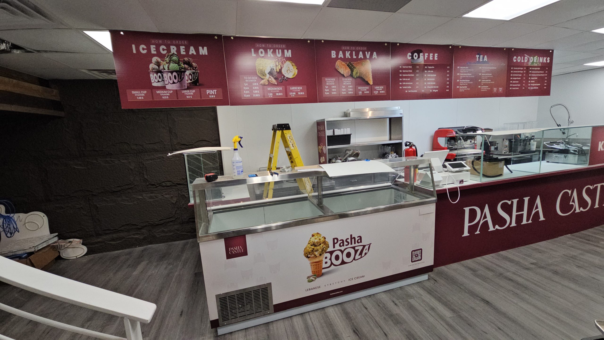 Interior of Pasha Castle ice cream shop with empty display freezer, a yellow ladder, coffee machine, and menu boards for ice cream, lokum, baklava, coffee, tea, and cold drinks.