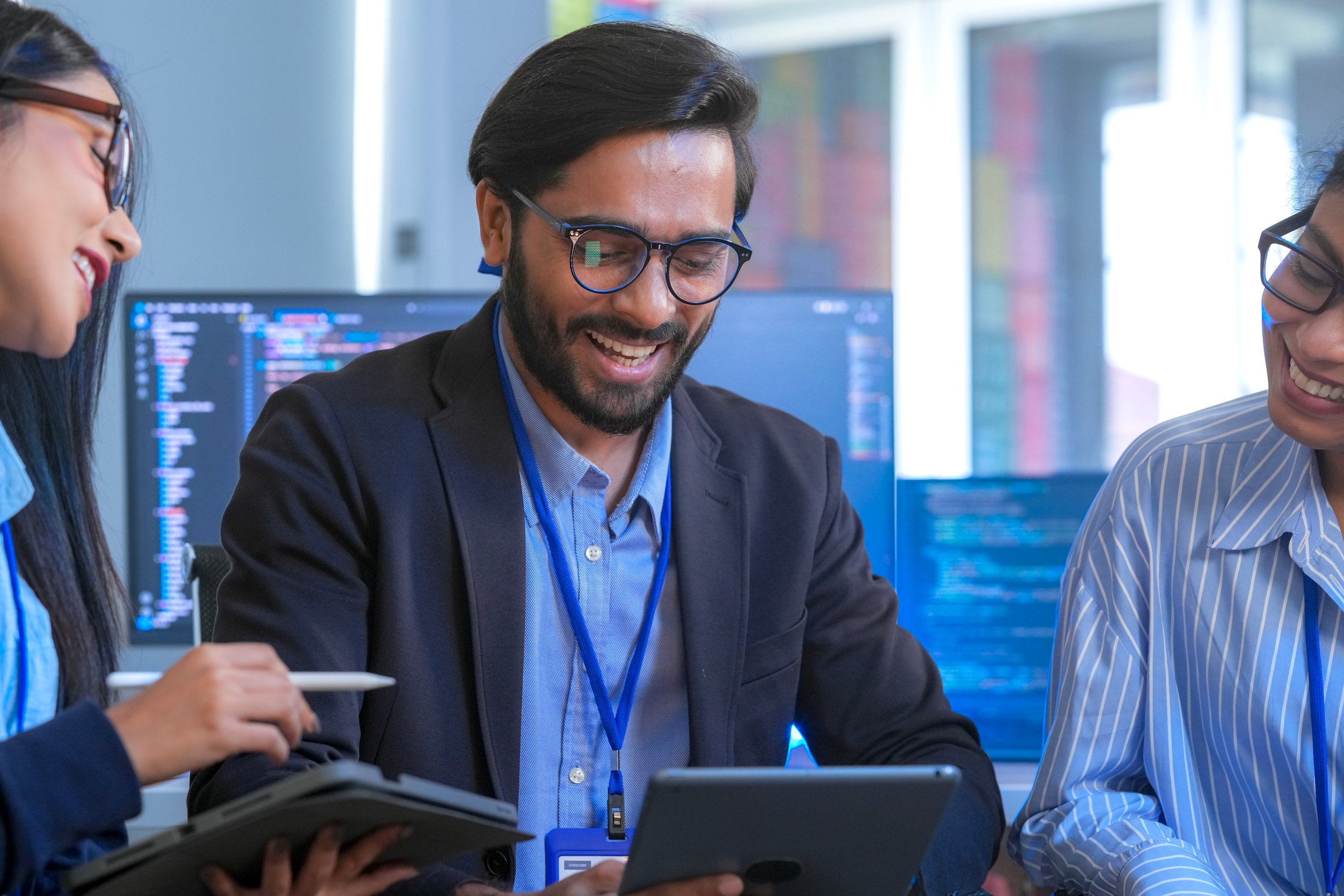 Three professionals smiling while discussing work using a tablet in an office with code on screens.
