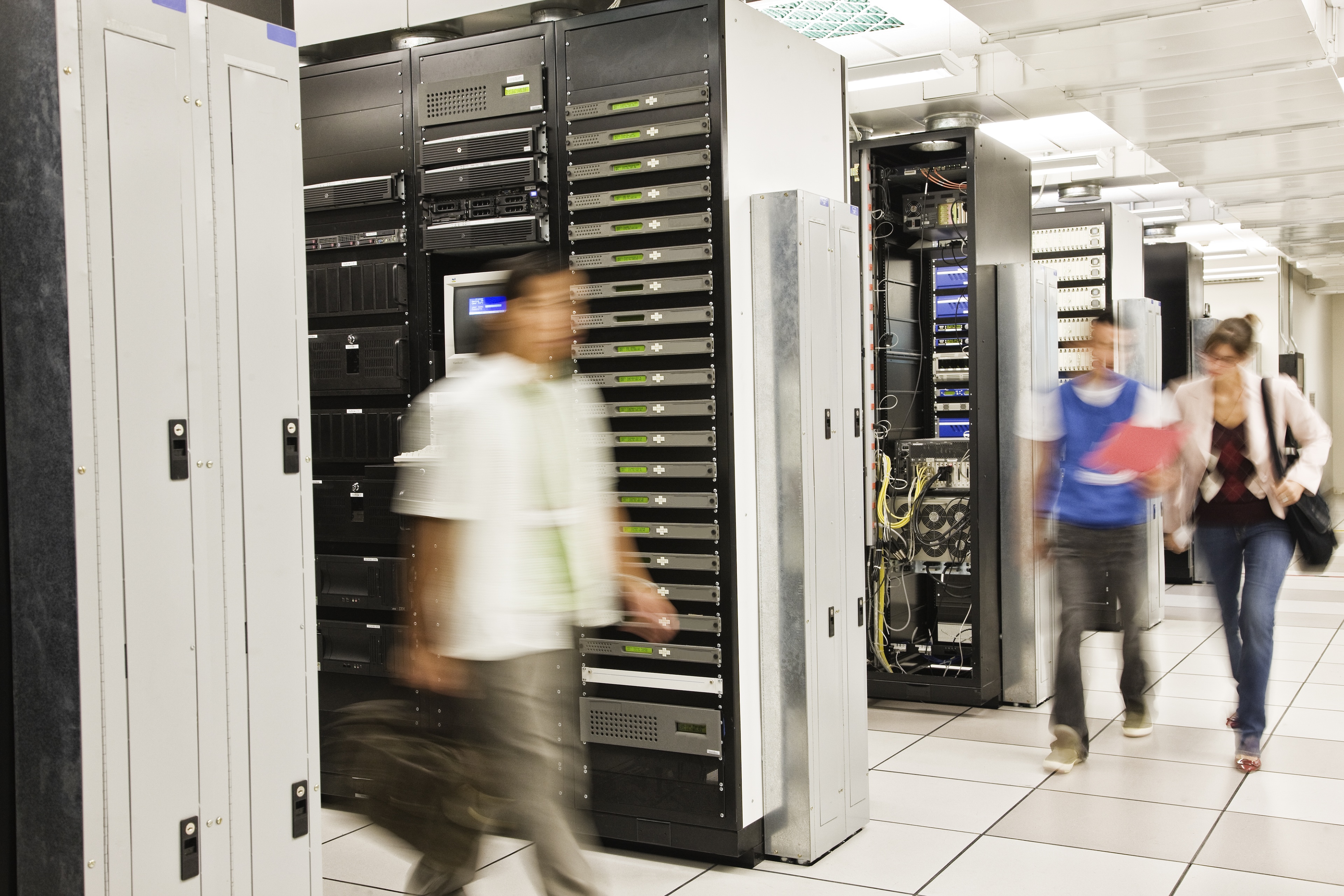 Blurred figures walking through a data center server room with racks of servers on both sides.