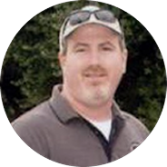 Man wearing a beige cap and sunglasses on his head with a dark polo shirt standing outdoors.