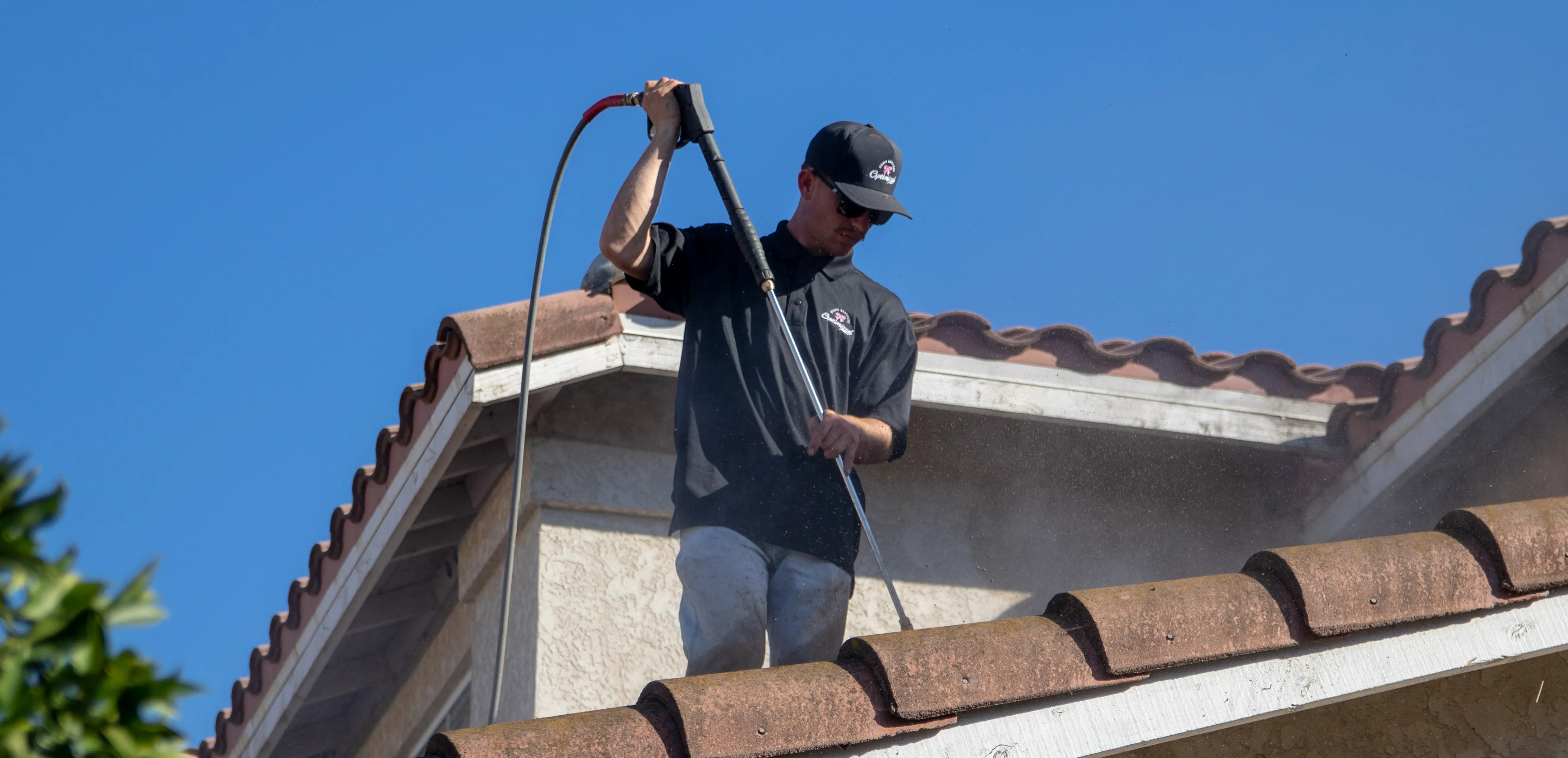 Man wearing a black cap and shirt pressure washing a tiled roof under clear blue sky.