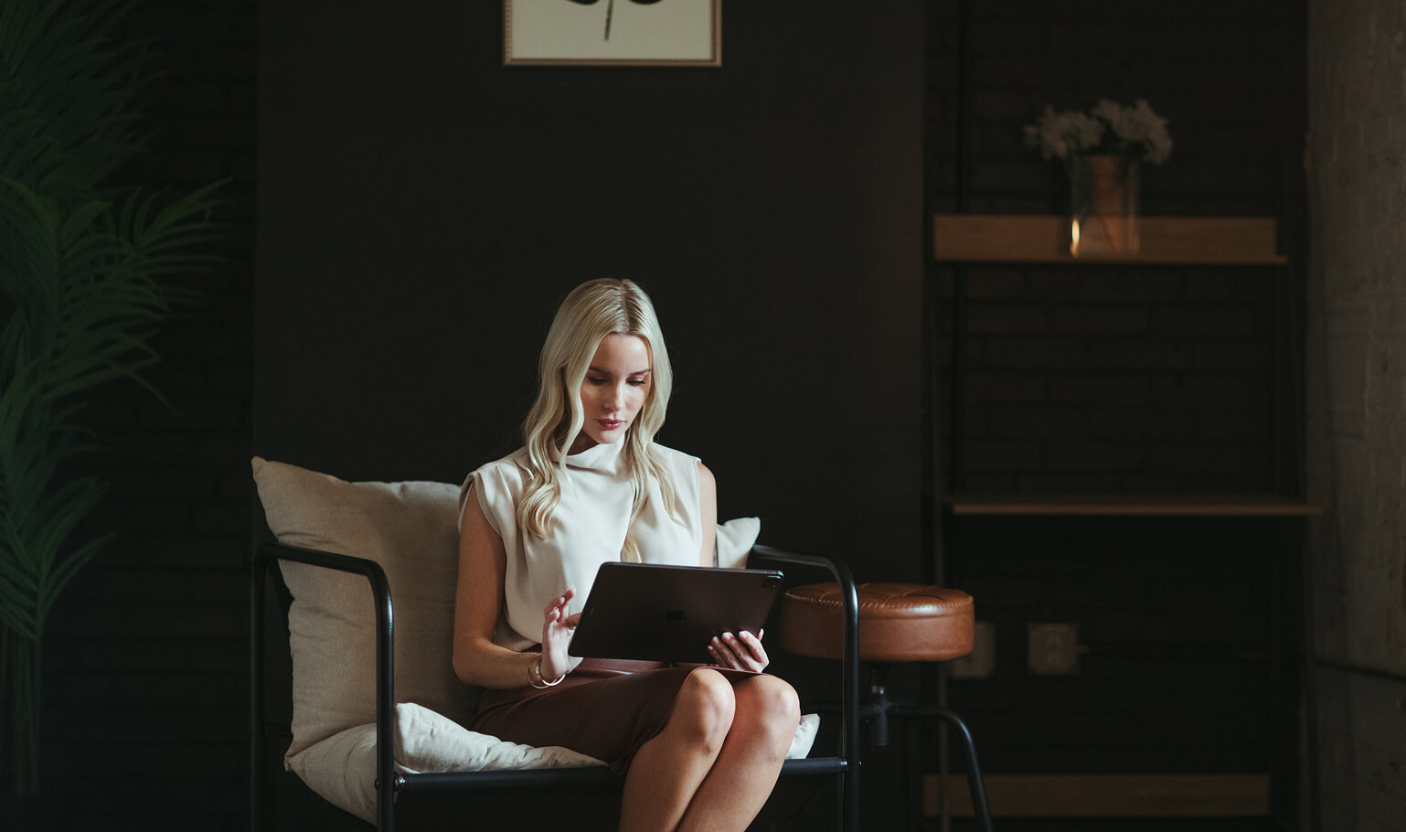 Blonde woman sitting on a cushioned chair in a dark room, using a tablet.