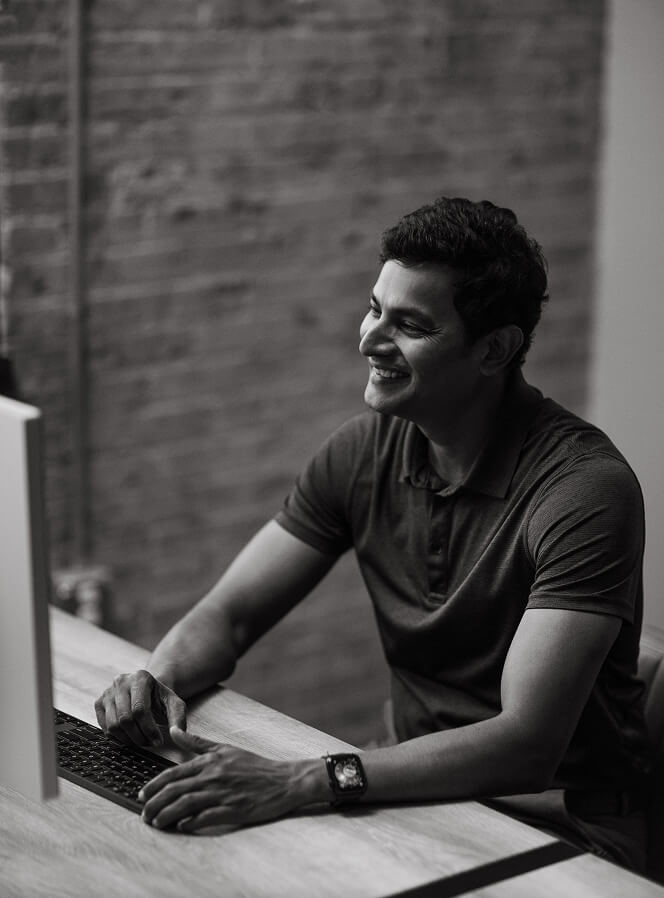 Smiling man sitting at a desk using a computer keyboard in a room with a brick wall background.