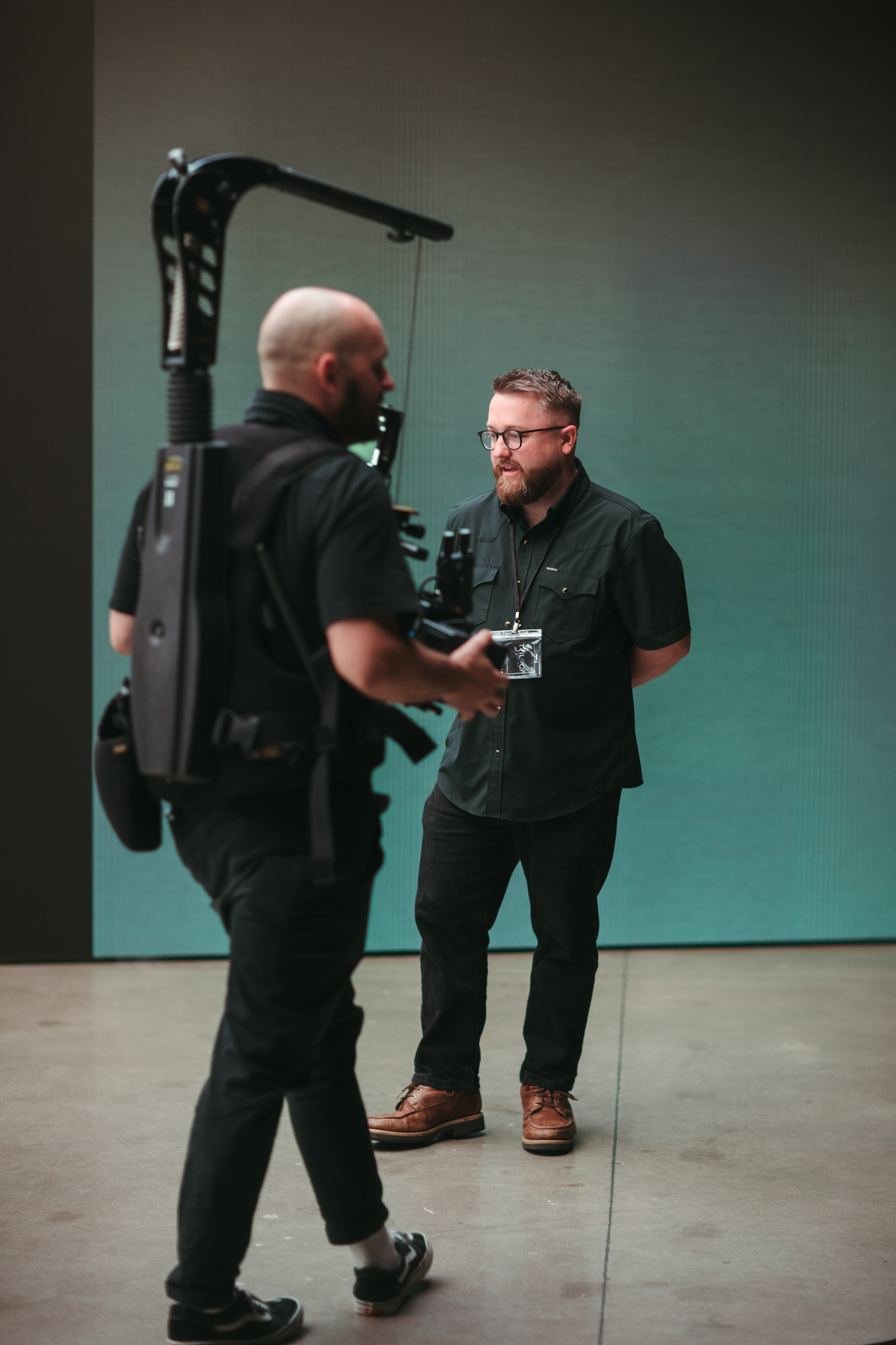 Photographer with camera rig filming a man in glasses and brown boots standing against a plain background.