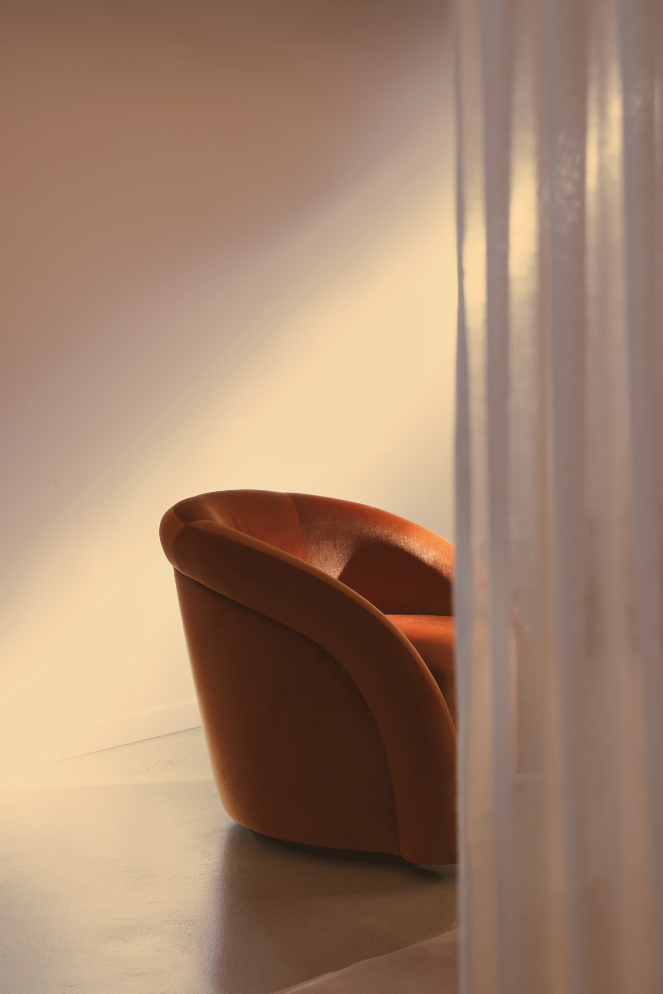 Curved brown armchair lit by soft warm light with sheer white curtain partially covering it.