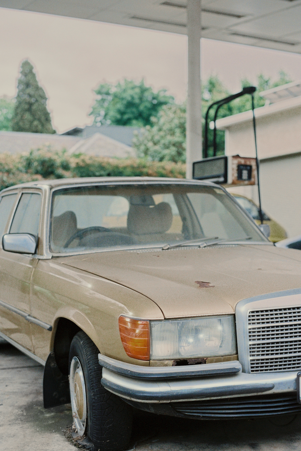 Old beige sedan with visible rust and flat front tire parked outdoors under a canopy.