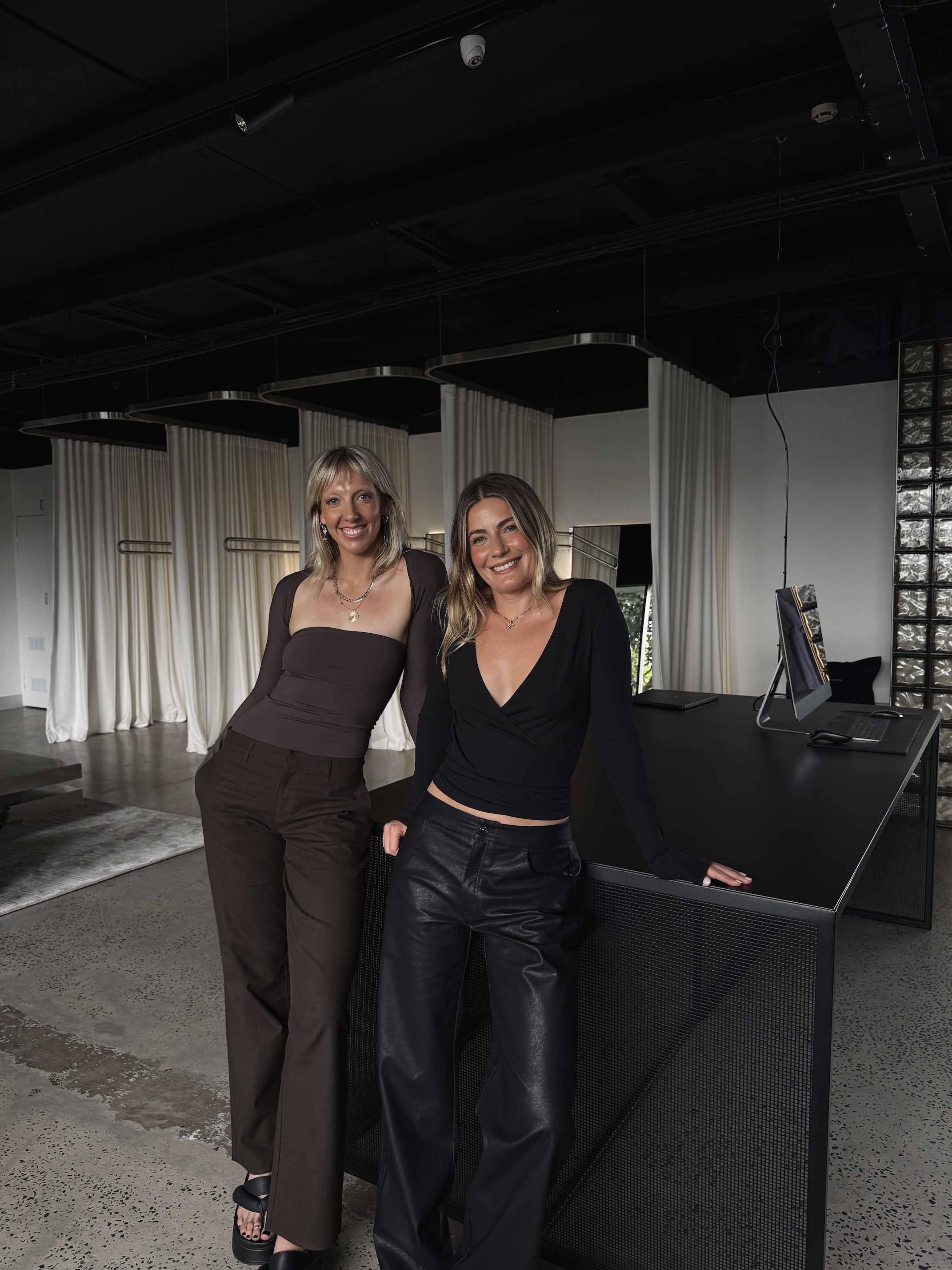 Two young women smiling, standing and leaning against a modern black desk in a stylish room with white curtains.