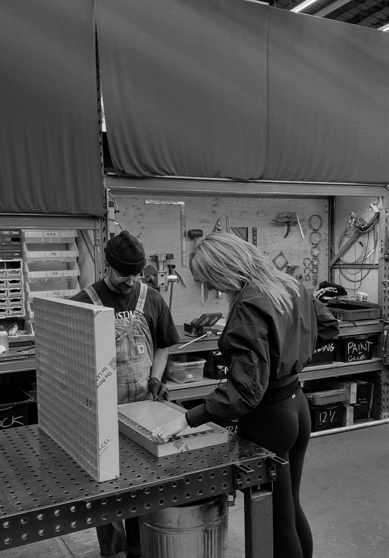 Two people in a workshop examining a rectangular object on a perforated metal table.