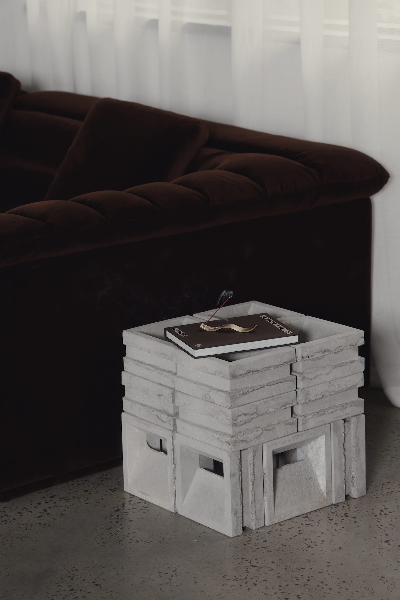 Concrete block side table next to a dark brown velvet sofa with a book and smoking incense stick on top.