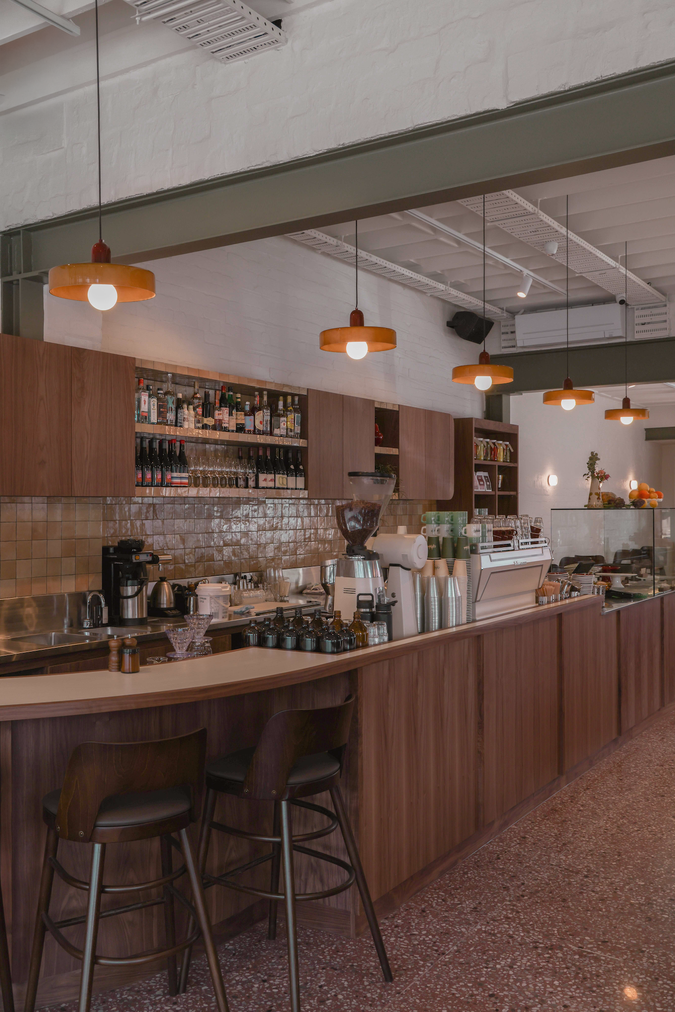 Modern coffee shop counter with wooden stools, pendant lights, coffee equipment, and bottled drinks displayed.