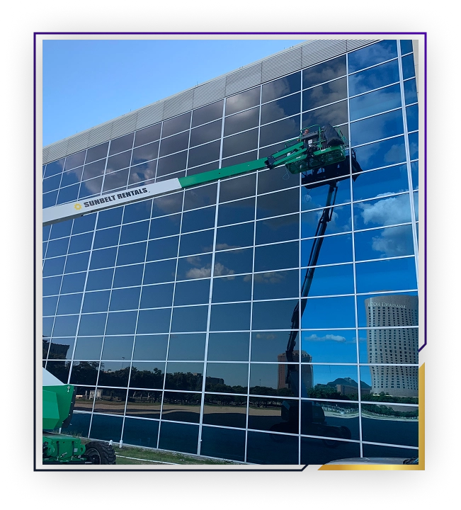 A green and white boom lift from Sunbelt Rentals extended to clean or work on the glass windows of a modern building reflecting a blue sky with clouds.