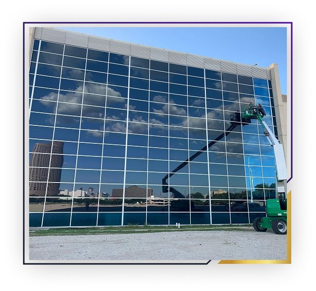 Worker in a green lift cleaning or maintaining the reflective glass windows of a modern building with blue sky and clouds reflected.