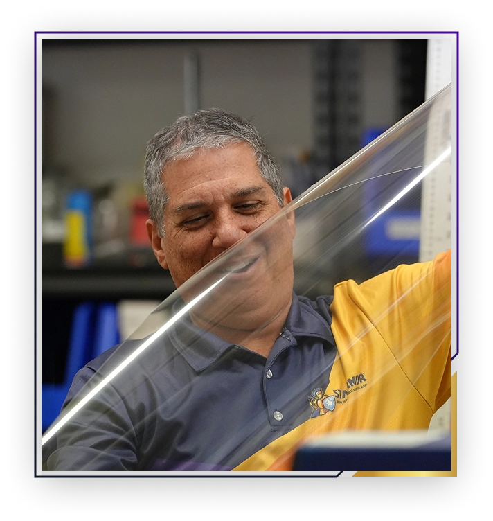 Man with gray hair and a yellow and navy polo shirt smiling while handling a clear plastic sheet in a workshop.