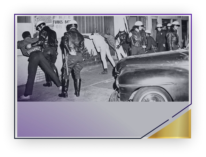 Black and white photo of police officers frisking multiple individuals against a wall during a street stop, with a car in the foreground.