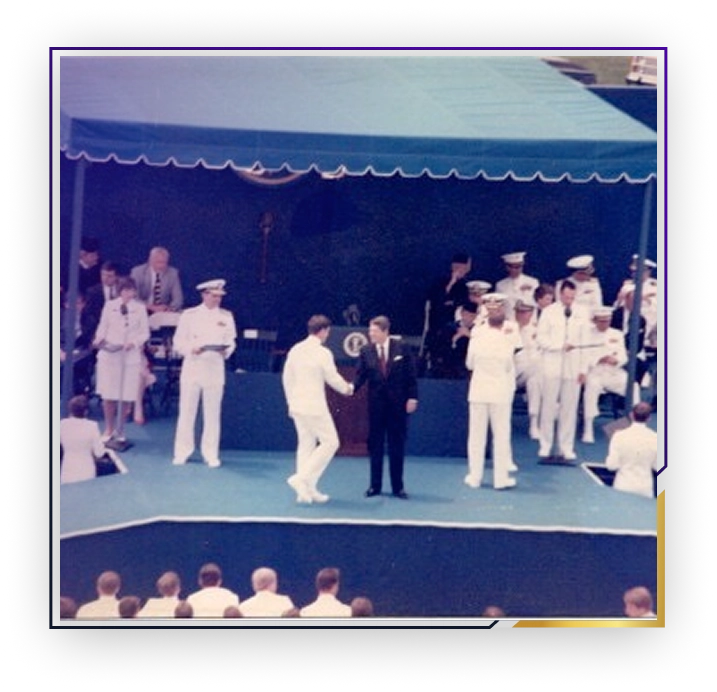 Person in white naval uniform shaking hands with a man in a dark suit on an outdoor stage with other naval officers and spectators present.