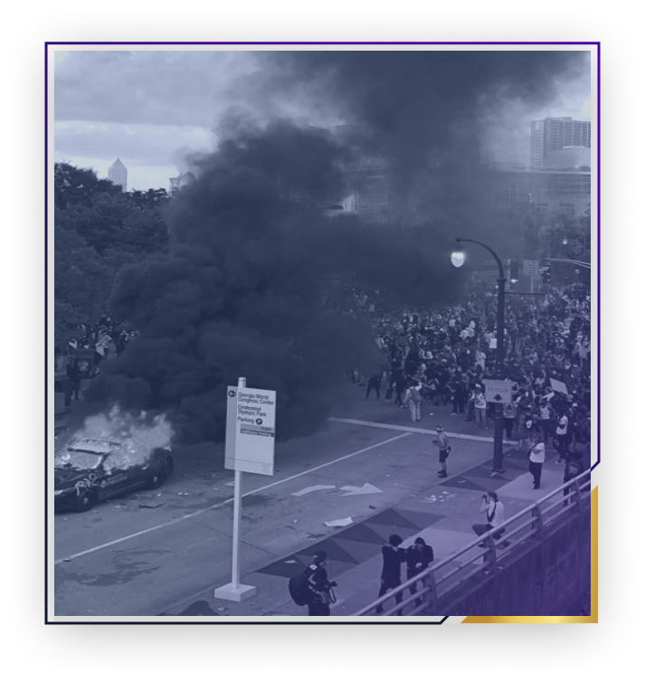 A crowd of people on a city street near a burning car emitting thick black smoke during a protest or riot.