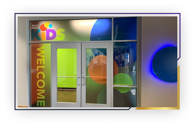 Entrance doors to Rock Creek Kids with colorful circular designs and green 'WELCOME' text on the left glass panel.