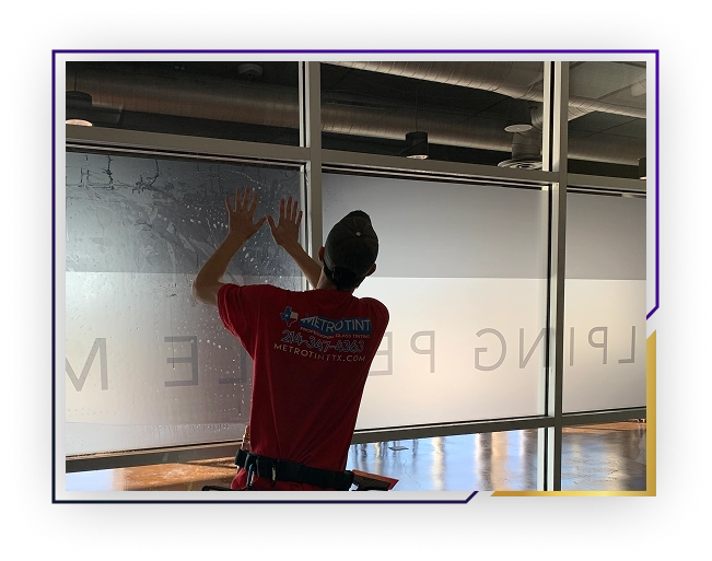 Person wearing a red Metro Tint shirt and cap installing a window film on a large glass window inside a commercial space.