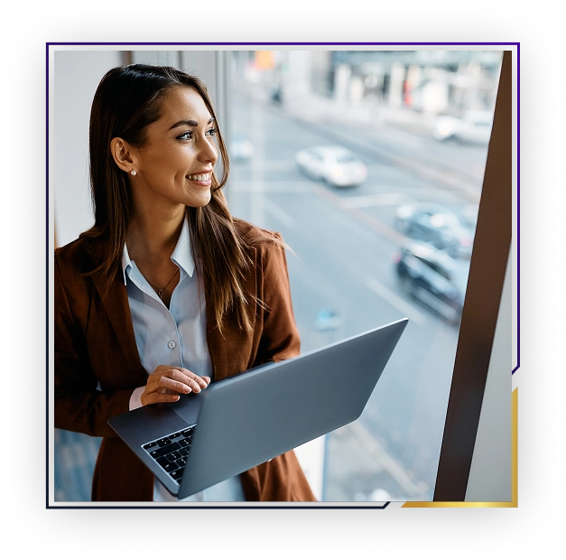 Smiling woman in brown blazer holding a laptop and looking out a window at a city street.