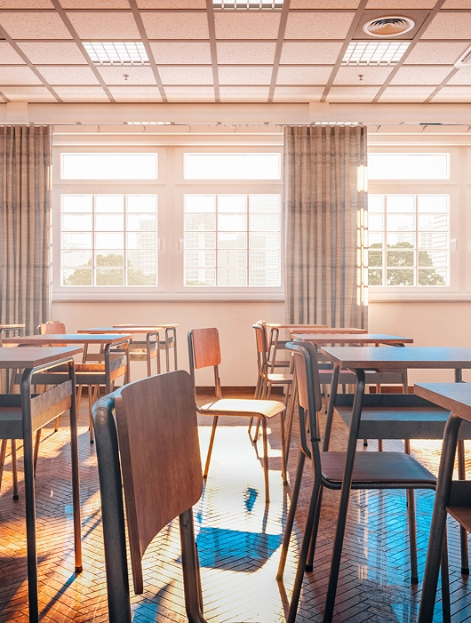 Sunlit empty classroom with wooden desks and chairs and large windows overlooking trees and buildings.