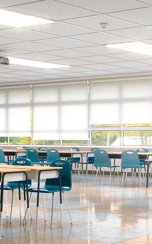 Empty classroom with rows of blue plastic chairs and wooden desks under fluorescent ceiling lights.