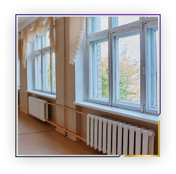 A school corridor with large windows and white radiators underneath, cream-colored walls, and beige curtains.
