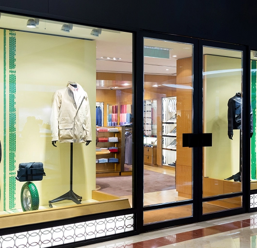 Clothing store window display with mannequins wearing beige and black jackets and shelves of folded clothes inside.