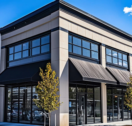 Corner view of a modern commercial building with large glass windows, black awnings, and a young tree in front under a blue sky.