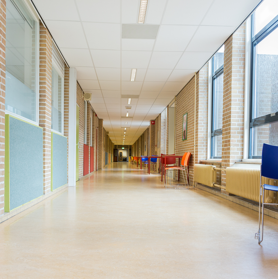 Long school hallway with brick walls, large windows on one side, and colorful chairs and tables along the corridor.