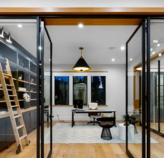Modern home office with glass double doors, wooden floor, a desk with computer, rolling chair, bookshelf with ladder, and a pendant lamp.