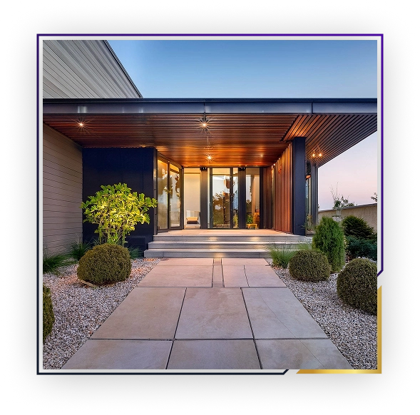 Modern house entrance with large tiled pathway, trimmed shrubs, a tree, and warm ceiling lights under a wooden canopy.